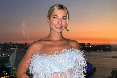 A woman in a feathered dress smiles, with a cityscape and sunset in the background.