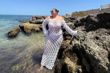 Woman in a white lace beach cover-up stands on rocks by the sea, under a clear blue sky.