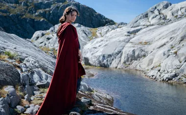 Character in red cloak stands by a rocky stream against mountainous backdrop in "Rings of Power".