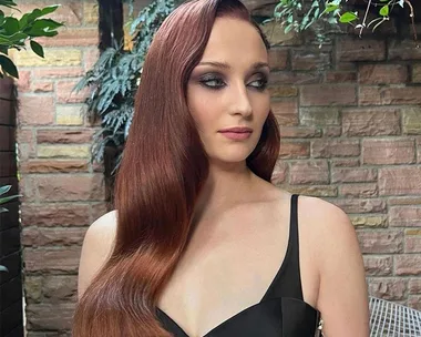 A woman with long red hair in a black gown stands against a brick wall and greenery, looking to the side.