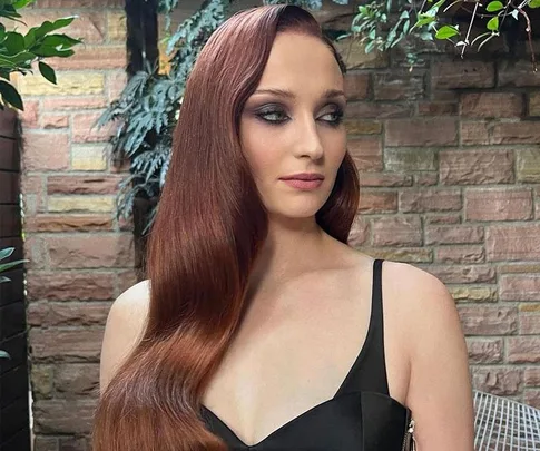 A woman with long red hair in a black gown stands against a brick wall and greenery, looking to the side.