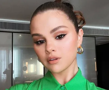 Portrait of a woman in a green shirt with bold eyeliner and hoop earrings, hair styled in a bun.