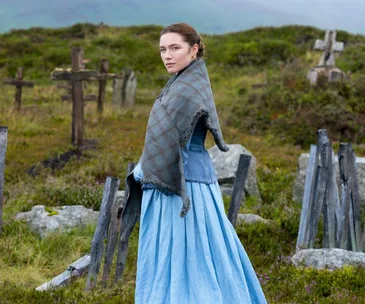 Woman in 19th-century attire stands in a graveyard, wearing a blue dress and plaid shawl, looking over her shoulder.