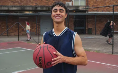 Young man smiling on a basketball court, holding a ball, wearing a sleeveless blue jersey.