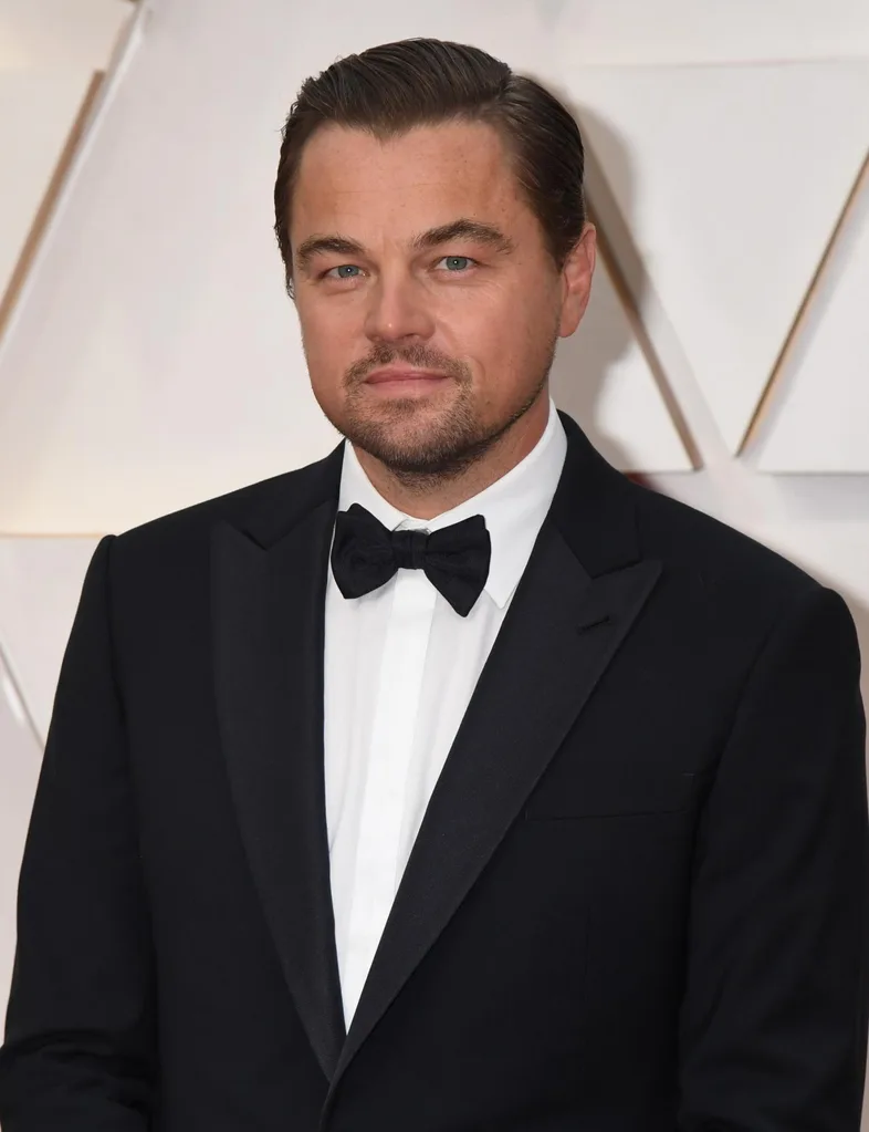 A man in a black tuxedo poses on a red carpet event backdrop.