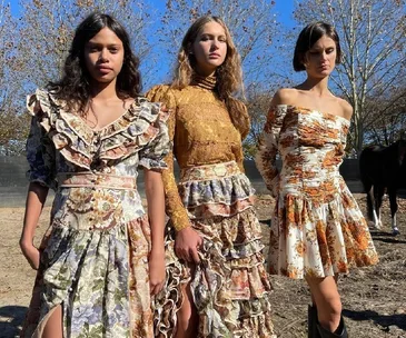 Three models in floral and lace dresses stand outdoors under clear blue sky.