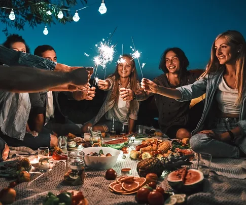 Group of friends at a nighttime picnic, holding sparklers and smiling, surrounded by food and lights.