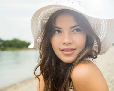 A woman in a white sun hat smiles, looking sideways. She has long brown hair and is outdoors near water.