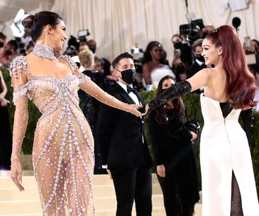 Two women in elaborate dresses smiling and holding hands at a high-profile event with photographers in the background.