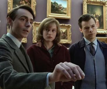 Three people stand in an art gallery, surrounded by framed paintings, engaged in a serious discussion.