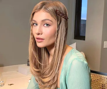 Young woman with long blonde hair and Gucci hair clip, wearing a light green top, posed indoors.