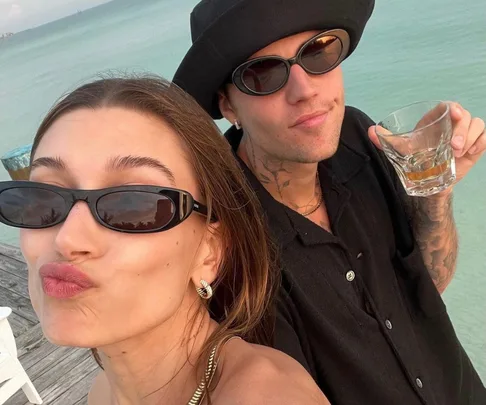 Two people in stylish sunglasses by the ocean, smiling, one holding a drink.