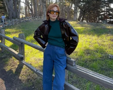 Person in sunglasses, leather jacket, and blue pants leaning on a wooden fence in a forest clearing.