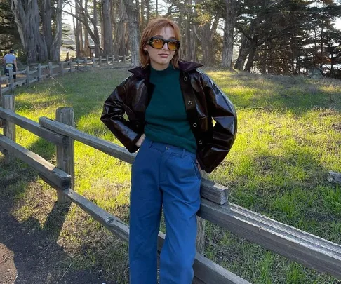 Person in sunglasses, leather jacket, and blue pants leaning on a wooden fence in a forest clearing.