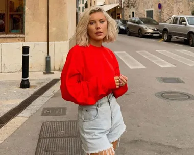 Woman with blonde hair in a red sweater and denim shorts standing on a street.