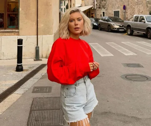 Woman with blonde hair in a red sweater and denim shorts standing on a street.