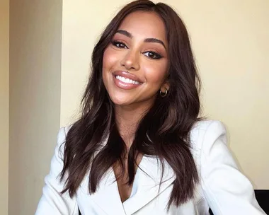 A woman with dark hair and hoop earrings smiles in a white blazer.