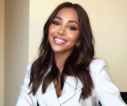 A woman with dark hair and hoop earrings smiles in a white blazer.