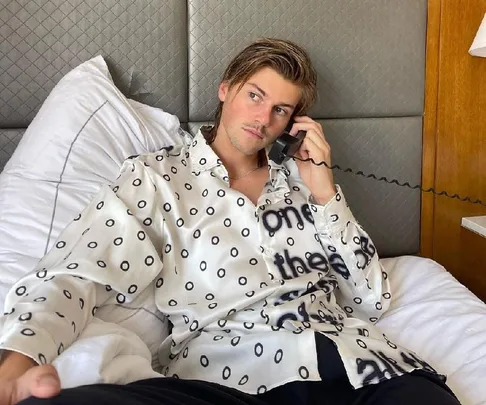 Person in a patterned shirt lies on a bed, holding a phone to their ear, looking pensive.