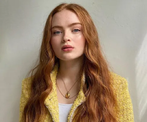 Young woman with long red hair wearing a yellow tweed jacket and necklace, posing against a plain background.