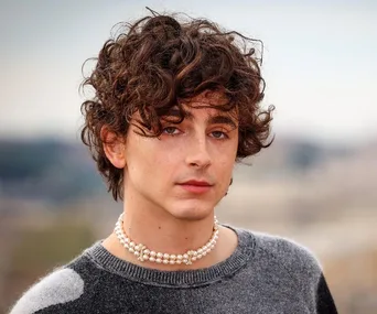 Young man with curly hair wearing a pearl necklace and a sweater, outdoors with a blurred cityscape background.