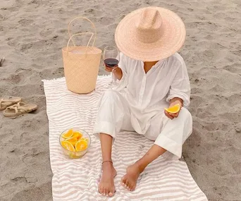 Person in white on a beach, seated on a striped towel with a sunhat, holding wine and orange slice, beside a woven bag.