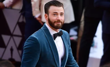 Man in a blue velvet suit with a beard and bow tie, smiling at an event with formal attire attendees in the background.