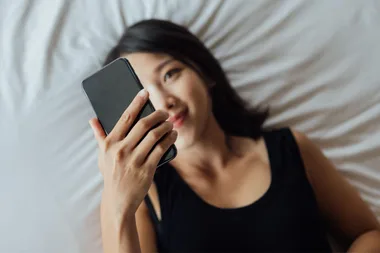 A woman in a black tank top lying on a bed, holding a smartphone and smiling.