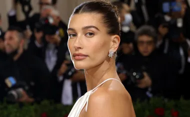 A woman with slicked-back hair and a white dress poses on the red carpet amidst photographers.