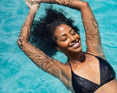 Woman smiling and floating in a pool, wearing a black bikini with sunlight reflecting on the water.