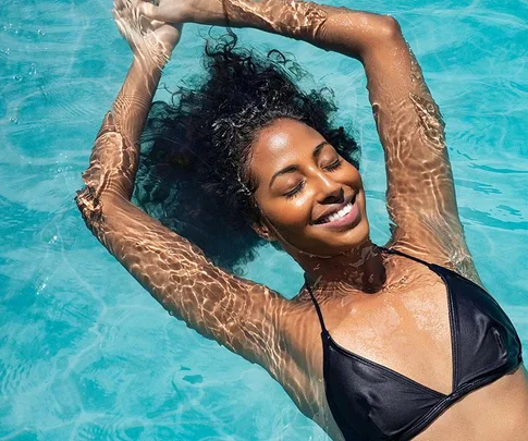 Woman smiling and floating in a pool, wearing a black bikini with sunlight reflecting on the water.