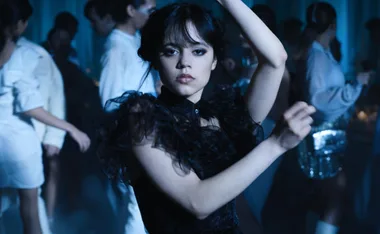 A young woman in a dark dress with lace details dances at a crowded event, surrounded by people in light clothing.