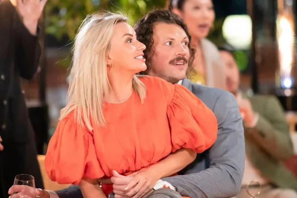 A blonde woman in an orange dress sits on a man's lap, both looking excitedly to their right.