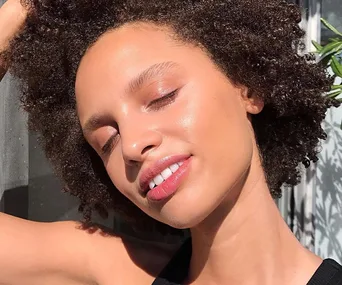 A woman with glowing skin and curly hair basks in sunlight, eyes closed, smiling contentedly.