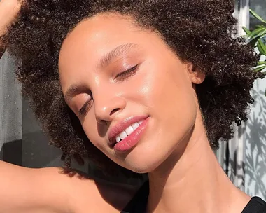 A woman with glowing skin and curly hair basks in sunlight, eyes closed, smiling contentedly.