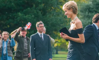 A woman in evening wear walks outdoors, warmly greeted by onlookers waving and taking photos.