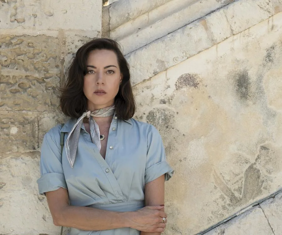 A woman in a light blue dress and scarf stands against a stone wall, arms crossed, gazing forward in a contemplative pose.