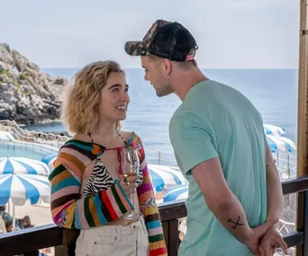 A woman with curly hair and colorful sweater holds a glass, smiling at a man in a green shirt, near the seaside.