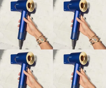 Hand holding a blue Dyson hair dryer with gold accents against a tiled background.