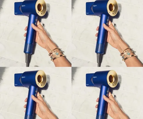 Hand holding a blue Dyson hair dryer with gold accents against a tiled background.