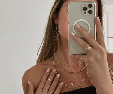 Woman taking selfie, wearing layered gold necklaces, earrings, rings, and holding a phone with a circular grip.