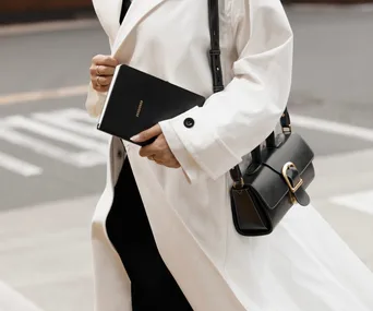 Person in white coat holding a black planner and handbag while walking on a city street.