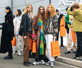 Group of stylish individuals with orange shopping bags, standing outside a store, dressed in various outerwear.