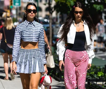 Two women in stylish outfits walking on a sunny city street; one in a checkered set, the other in a black top and red pants.