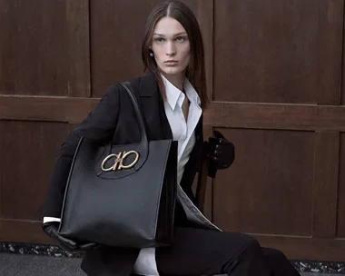 Woman in black jacket holds a large black tote bag with gold emblem, sitting against a wooden backdrop.