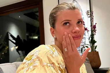 Woman showing off an engagement ring while sitting indoors, wearing a yellow floral top.