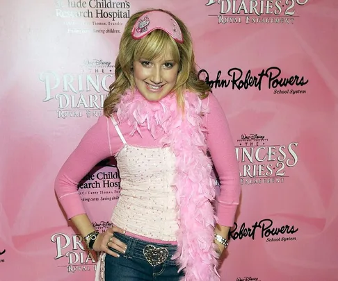 A woman in a pink outfit with a feather boa poses at a "The Princess Diaries 2: Royal Engagement" event backdrop.