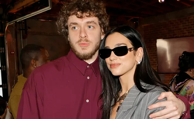 A man in a maroon shirt poses with a woman in sunglasses and a pinstripe blazer, with a hand on her shoulder.