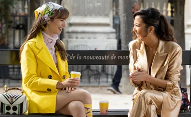 Two women sit on a bench in Paris, one in a yellow coat, the other in a gold suit, both smiling and holding coffee.