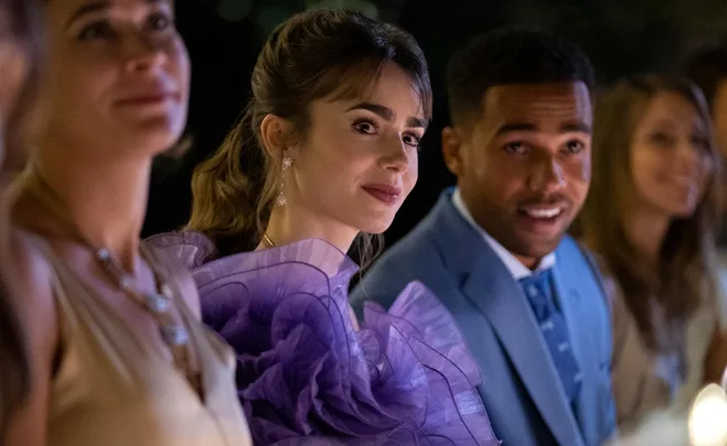 Emily in a ruffled purple dress sits at a dining table, smiling, surrounded by other guests.